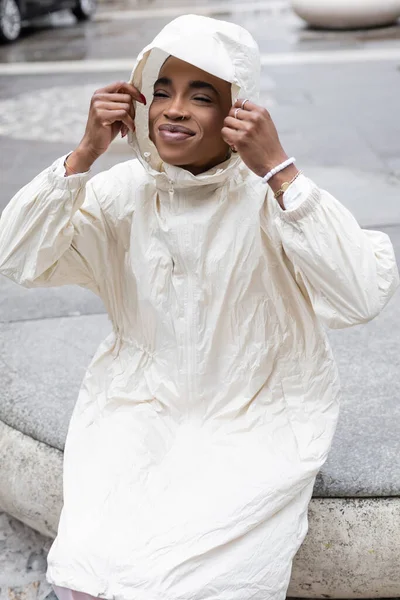 Jovem afro-americana de capa de chuva sentada na rua urbana turva — Fotografia de Stock