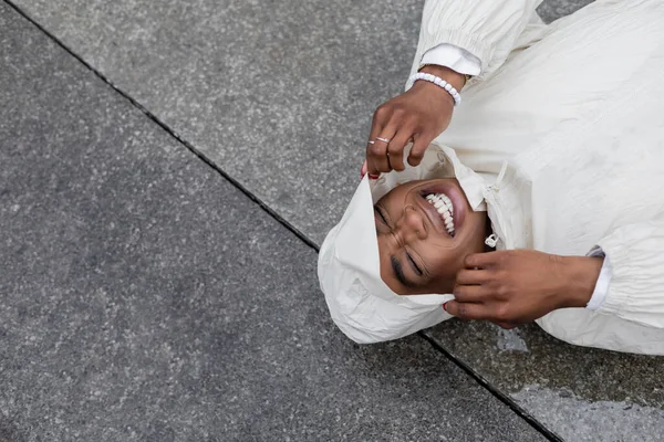 Top view of cheerful african american woman in raincoat lying on urban street — Stock Photo