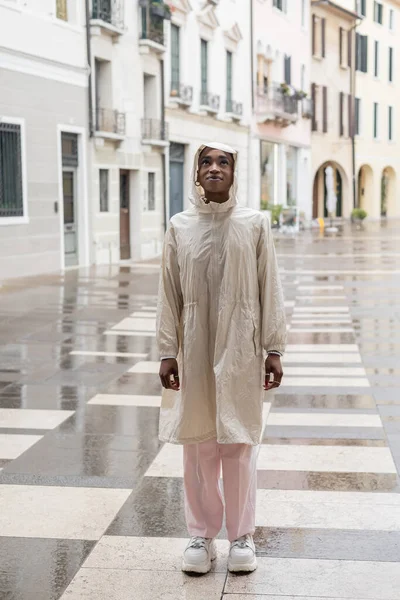 Fashionable african american woman in raincoat standing on blurred street in Italy — Stock Photo