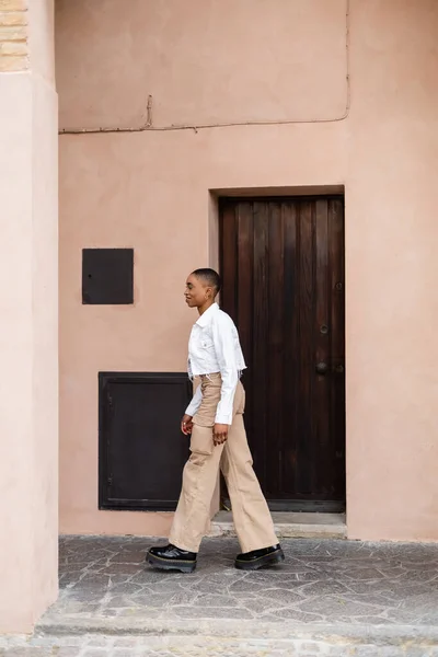 Vista lateral de la sonriente mujer afroamericana caminando cerca del edificio en la calle en Italia - foto de stock