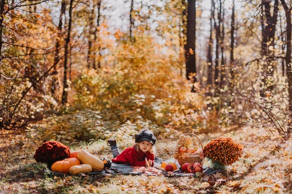 Little girl in a red coat sits in an autumn forest with a basket of autumn fruits