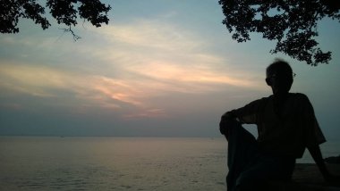 Ağacın altında rahatlayan yaşlı bir adamın silueti. Arkaplan olarak renkli gün batımı gökyüzü ve nehir