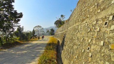 Üç kimliği belirsiz kişi eğimli eğimli yamaçlı yolda yürüyor, arka planda tepeler var. Sağdaki beton destek duvarı.
