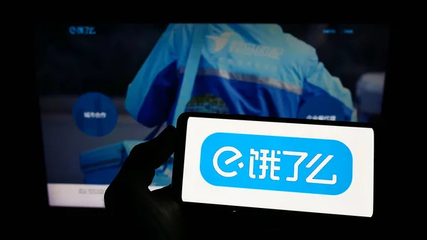 Person holding mobile phone with logo of Chinese food delivery company ELEME Inc. (ele.me) on screen in front of web page. Focus on phone display.