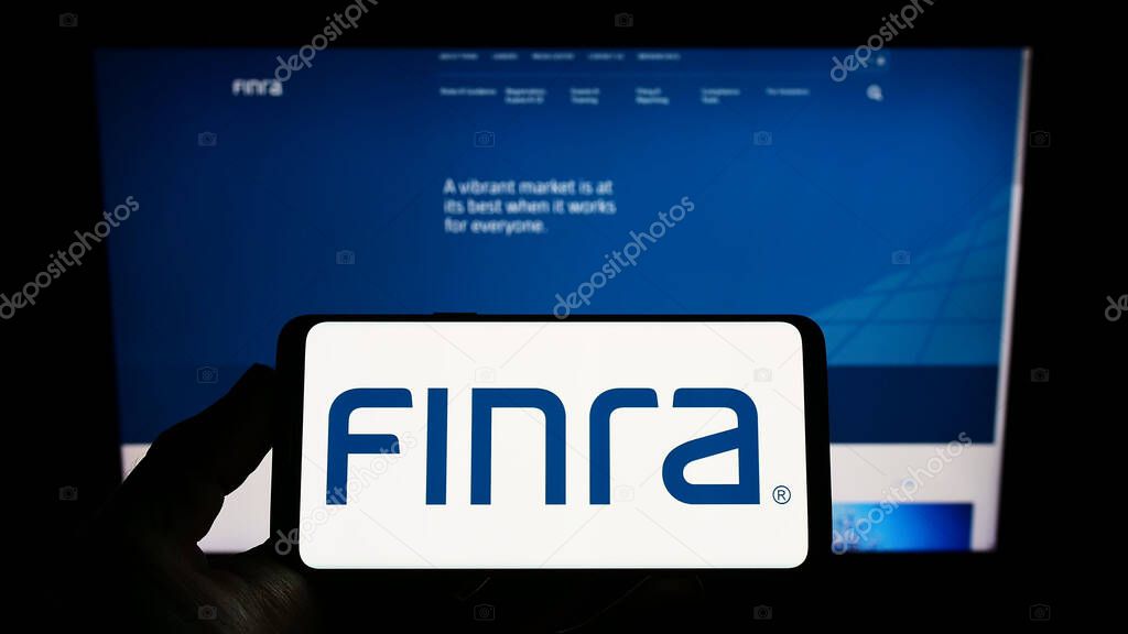 Person holding cellphone with logo of Financial Industry Regulatory Authority (FINRA) on screen in front of webpage. Focus on phone display.