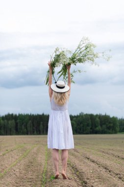 Güzel sarışın bir kadının dikey bir fotoğrafı. Şapka takıyor ve elinde çiçekleri tutan beyaz bir elbise.