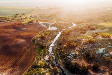 Gün batımında gökyüzünün altında otlakların, çalıların ve su kanallarının havadan çekilmiş görüntüsü.