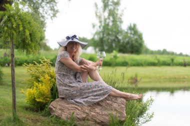 Güzel sarışın bir kadın elinde şarap kadehi ve yeşil bir tarladaki bir gölün yanındaki kayanın üzerinde oturuyor.