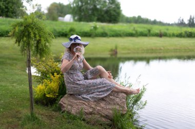 Güzel sarışın bir kadın elinde şarap kadehi ve yeşil bir tarladaki bir gölün yanındaki kayanın üzerinde oturuyor.
