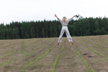 Arkasında yeşil ağaçlar olan bir tarlada zıplayan güzel sarışın bir kadın.
