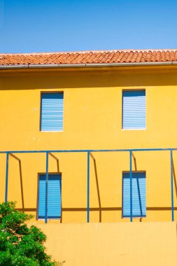 Mavi gökyüzüne karşı fayanslı çatılı sarı binanın ön cephesi.