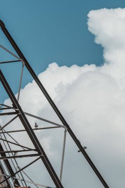 Mavi bulutlu arka planda demir vinç yapısında oturan iki güvercinin dikey görüntüsü.
