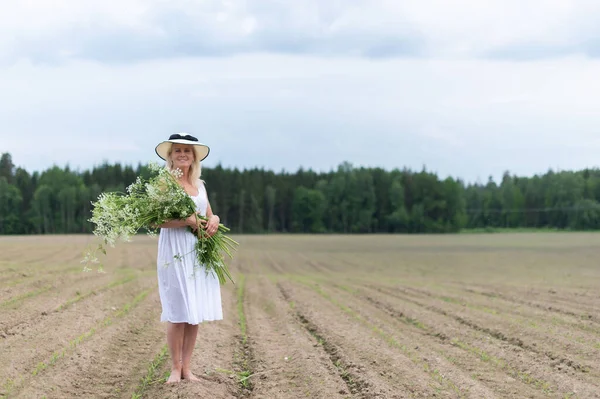 Güzel sarışın bir kadın, şapkalı ve beyaz elbiseli, elinde çiçeklerle tarlada.