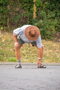 Deri şapkalı ve şortlu yaşlı bir adamın dikey görüntüsü. Sokaktan bir şey almak için eğiliyor.
