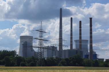 Almanya 'nın Hanau kenti yakınlarındaki Staudinger kömür yakıtlı enerji santrali, yüksek voltajlı hatlarla bir tahıl tarlasının arkasında mavi-beyaz gökyüzüne karşı.