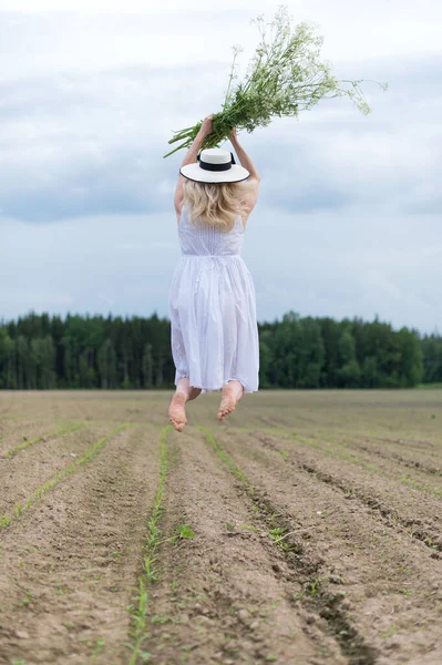 Beyaz elbiseli güzel bir kadının elinde çiçeklerle bir tarlada zıpladığı dikey bir fotoğraf.