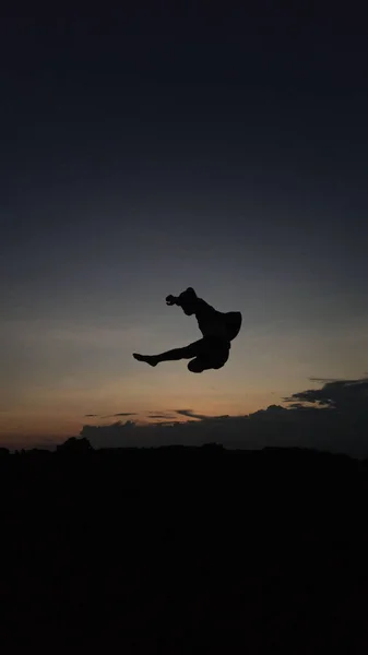 A vertical silhouette of a male gymnast doing a somersault in sunset
