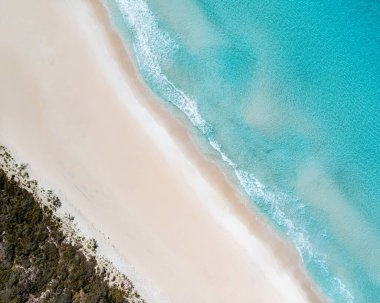 Gökyüzü kumlu bir plajın ve kristal berrak, deniz mavisi bir suyun hava aracı görüntüsü.