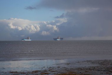 Bulutlu bir gökyüzünde seyreden iki büyük gemiyle sakin bir sahil.