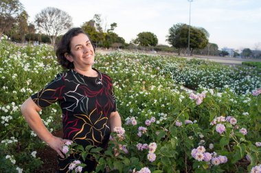 Güneşli bir günde parkta olgun bir kadın. Bir çiçek tarhında dikilip eğleniyor.
