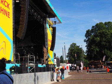 Güneşli bir günde elinde müzik aletleriyle ana sahnenin yanında duran insanların görüntüsü.