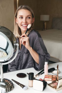 Güzel sarışın bir kadın makyaj fırçasıyla yüzünü kızartıyor, gülümsüyor ve aynaya bakıyor. Elit kozmetik ürünleri