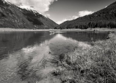 Kurumuş çimenlerin arasından bulutların yansımaları ve uzak karlı dağlarla kaplı temiz bir göl manzarası..