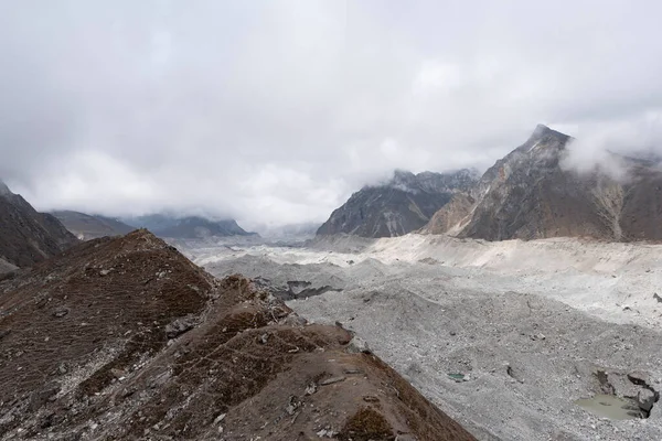 Bulutlu arka planda dağlarla çevrili kumlu bir alanın manzarası.