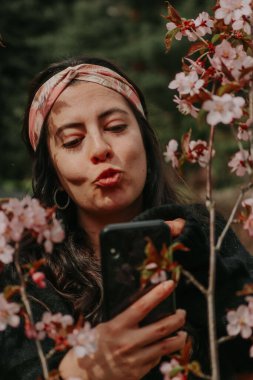 Parkta telefonuyla selfie çeken İspanyol bir kızın portresinin dikey görüntüsü.