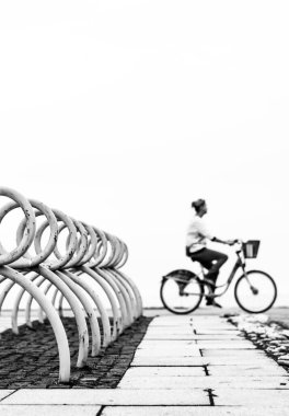 Bisiklet parkının yakınındaki bisiklete binen bir kadının dikey gri tondaki görüntüsü.