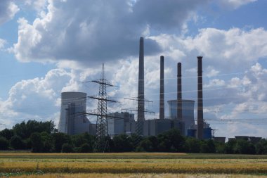 Almanya 'nın Hanau kenti yakınlarındaki Staudinger kömür yakıtlı enerji santrali, yüksek voltajlı hatlarla bir tahıl tarlasının arkasında mavi-beyaz gökyüzüne karşı.