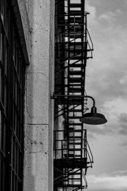 Duvarda asılı bir lambanın dikey gri tonda görüntüsü ve arka planda yangın merdiveni.