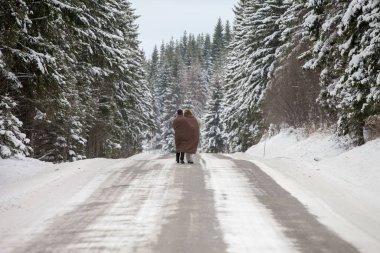 Karla kaplı çam ağaçlarıyla çevrili yolda battaniyeye sarılı kalan çiftlerin arka planı.