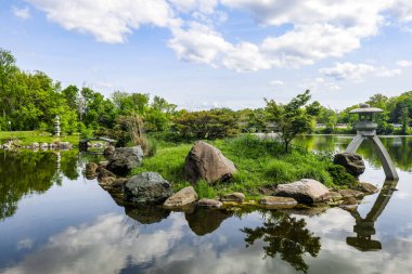 Göllü, otlu ve ağaçlı güzel bir park. Arkasında bulutlu mavi bir gökyüzü var.