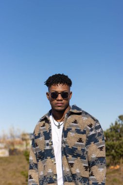 Mavi gökyüzünün altında kısa Afro saçlı ve güneş gözlüklü genç bir İspanyol 'un dikey görüntüsü.