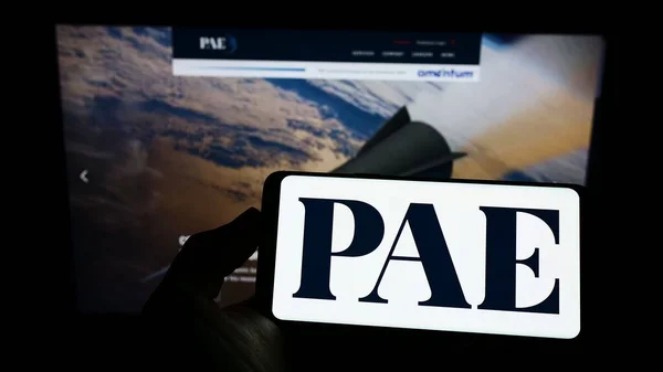 Person holding cellphone with logo of US company Pacific Architects and Engineers LLC (PAE) on screen in front of webpage. Focus on phone display.
