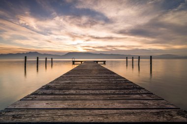 Günbatımı bulutlu gökyüzü ile Avusturya Attersee Gölü 'ne giden ahşap bir iskelenin büyüleyici bir görüntüsü.
