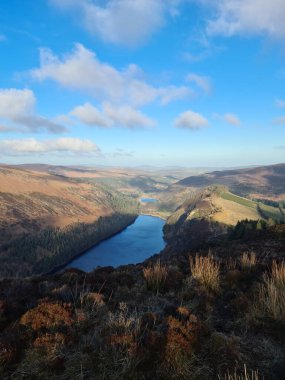 Güneşli bir günde, bulutlu mavi gökyüzünün altında yeşil tepelerle çevrili bir gölün yüksek açılı görüntüsü.