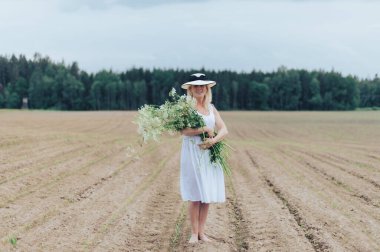 Güzel sarışın bir kadın, şapkalı ve beyaz elbiseli, elinde çiçeklerle tarlada.