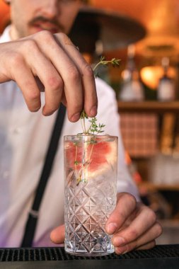 Barmenin barın üzerine şeffaf bir kokteyl bardağına garnitür koyduğu dikey bir fotoğraf.