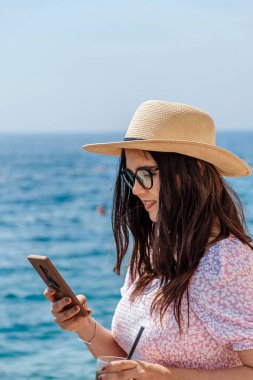 Kafkasyalı beyaz bir kızın bulanık bir deniz arka planında telefonuna baktığı dikey bir fotoğraf.