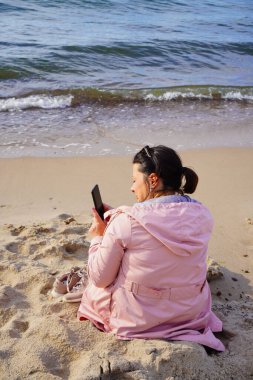 Pembe yağmurluk giymiş orta yaşlı bir kadının güneşli bir plajın fotoğraflarını çekerken çekilmiş dikey bir fotoğrafı.