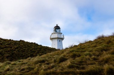 Bulutlu mavi gökyüzüne karşı iki yeşil tepe arasında beyaz bir deniz fenerinin manzarası