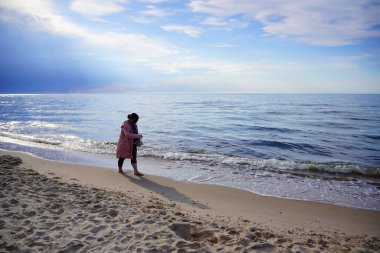 Pembe yağmurluk giymiş orta yaşlı tombul bir kadın güneşli bir sahilde yalınayak yürüyor.