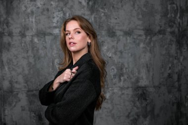 Gri bir duvarın önünde poz veren zarif, koyu renk elbiseli beyaz bir kadın.