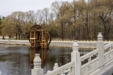 Beyaz bir köprünün yanındaki göldeki ahşap bir su tekerleğinin ve arka plandaki bir ormanın güzel bir görüntüsü.