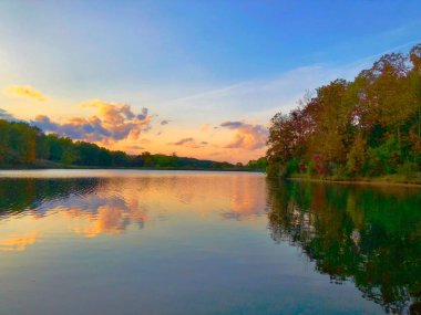 Sonbaharda renkli ağaçların arasından akan nehirde yansıtılan nefes kesici bulutlu bir gün batımı.