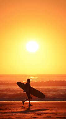 Sahilde yürüyen ve gün batımında dalgalara binmek için sörf tahtasını taşıyan bir adamın silüeti.