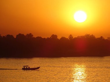 Sakin bir sahilde, ormanın siluetine ve parlak günbatımına karşı seyreden küçük bir tekne.