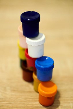 Ahşap bir masada, arka planda bulanık bir şekilde duran, çok renkli, küçük suluboya kaplardan oluşan bir yığının yakın plan çekimi.